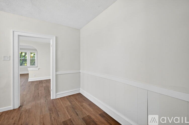 A room with white walls and wooden flooring, leading to a hallway with a window.
