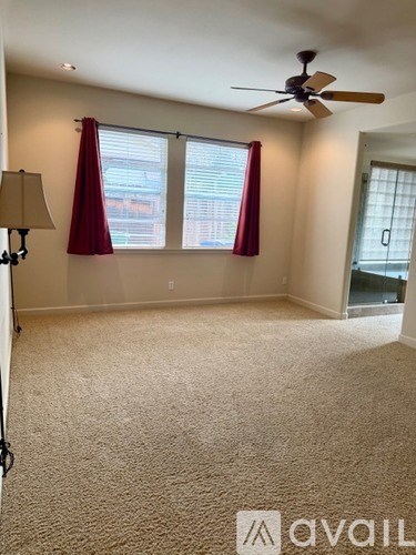 A room with a carpeted floor, a ceiling fan, and a window with red curtains.