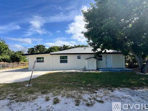 A white house with a grey roof is surrounded by greenery and has a clear blue sky above it.