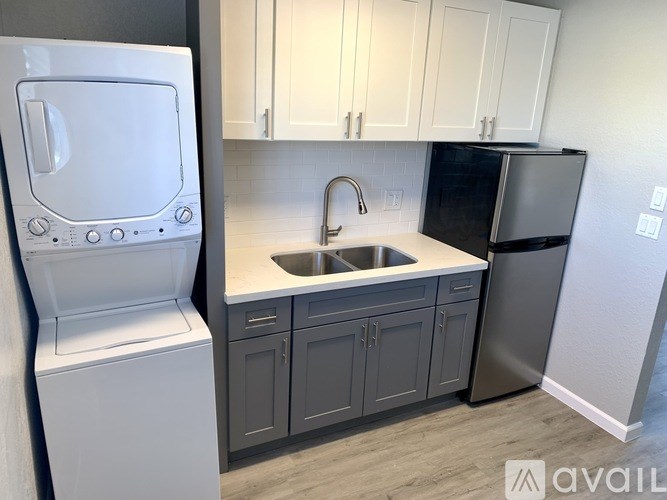 A white washing machine is in the foreground of a kitchen with a sink and a black refrigerator.