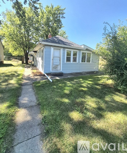 A small house with a grey roof and a white door is surrounded by grass and trees.