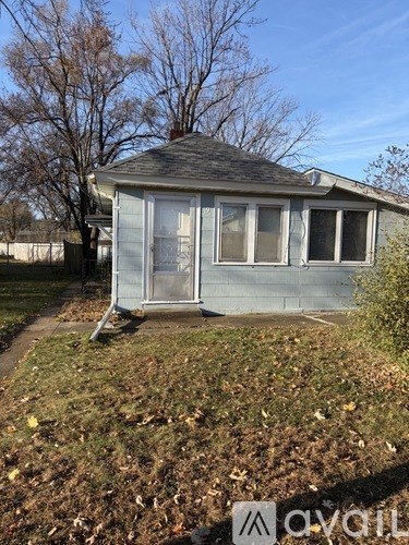 A small house with a grey roof and a white door is surrounded by grass and trees.