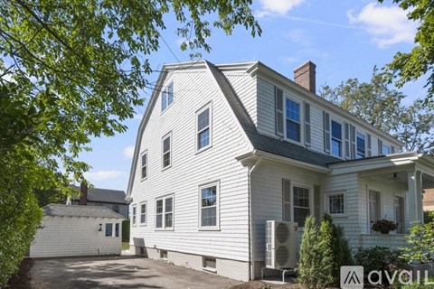 A white house with a grey roof and a chimney is surrounded by greenery.