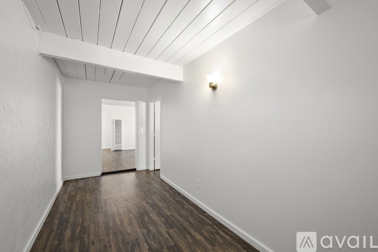 A room with wooden floors and white walls, with a light on the wall and a doorway in the distance.