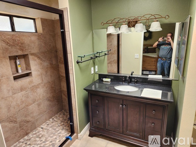 A man is taking a picture of a bathroom with a sink, mirror, and shower.