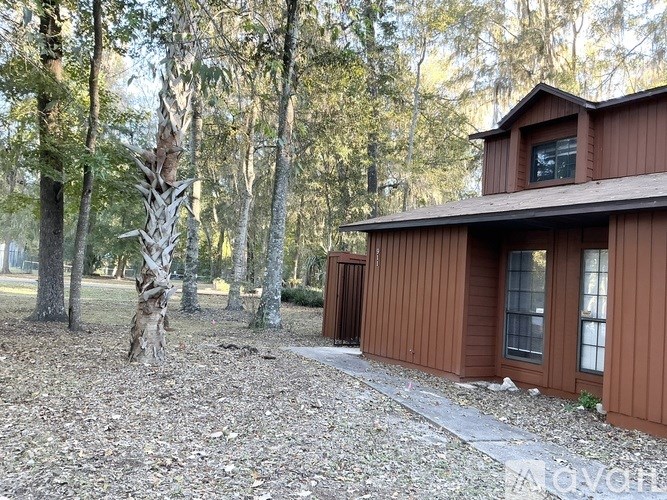 A house with a brown siding is surrounded by trees and has a gravel driveway.