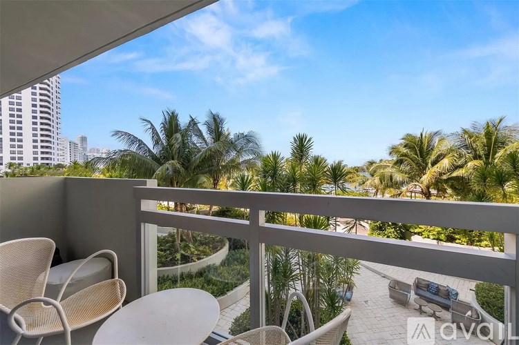 A balcony with chairs and a table overlooks a tropical garden and buildings.