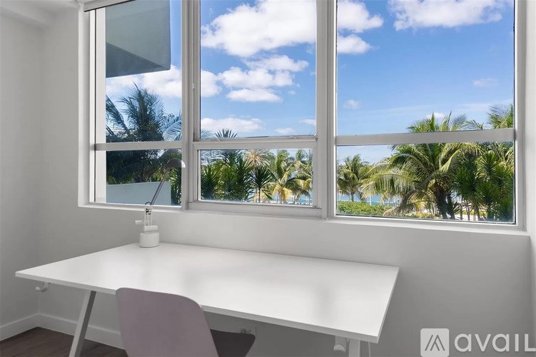 A white desk with a chair in front of a window with a view of a palm tree lined beach.
