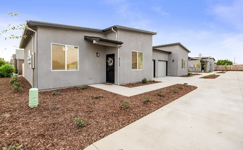 A grey house with a black door and windows is surrounded by a gravel area and a concrete pathway.