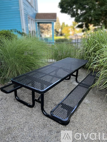 A black metal picnic table is in the foreground with a blue building and greenery in the background.