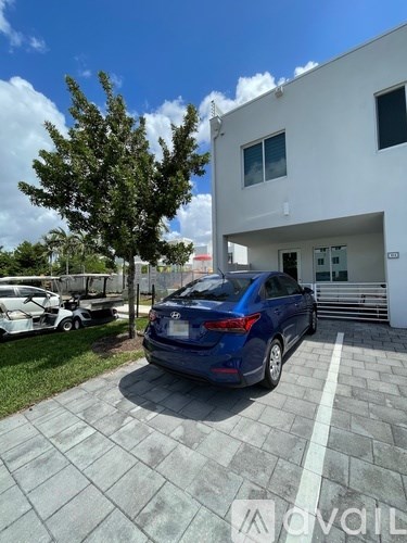 A blue car is parked in front of a white building with a tree to the left.