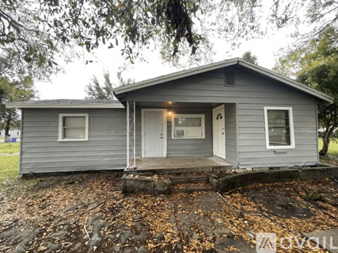 A grey house with a white door and windows is surrounded by fallen leaves.