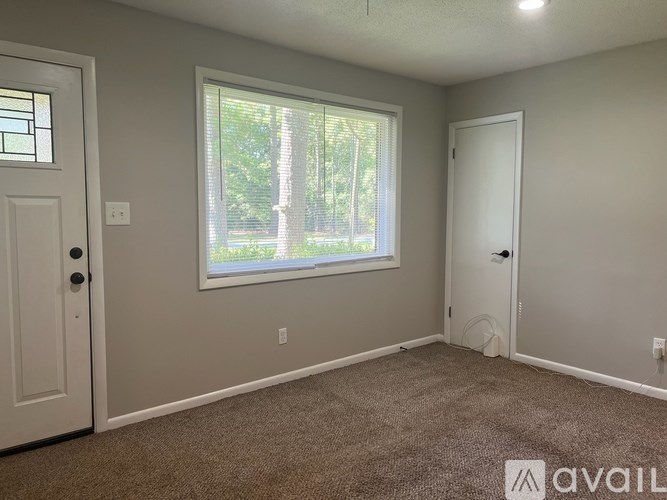 A room with a carpeted floor, a window with a view of trees, and a door on the left.
