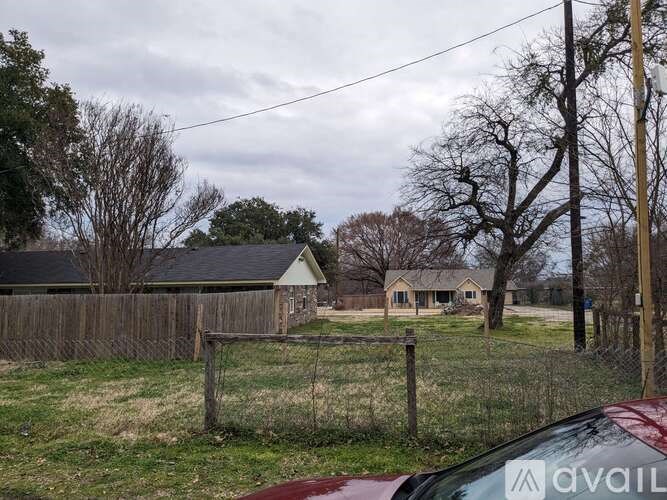 A red car is parked in front of a house with a fence and trees in the background.