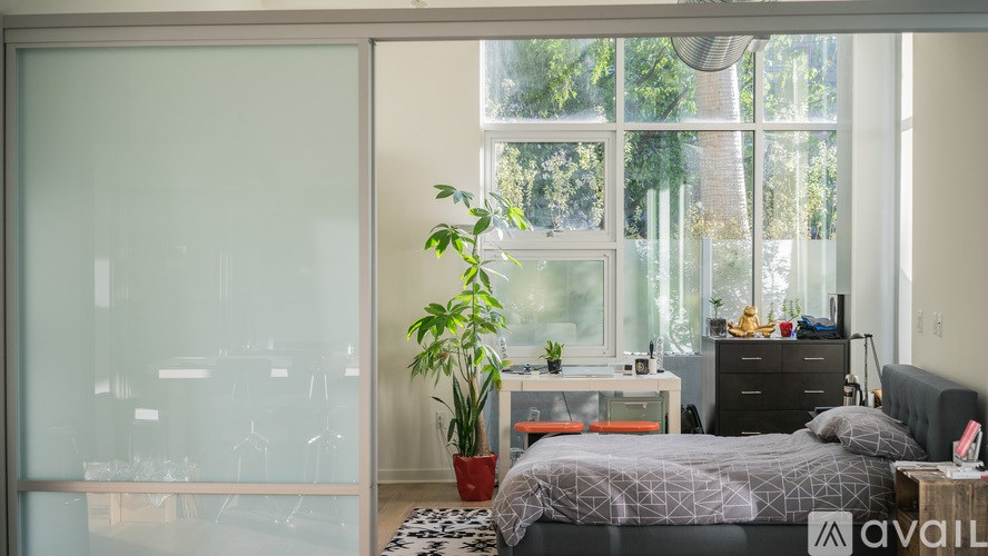 A bedroom with a bed, nightstand, and a plant in front of a large window.