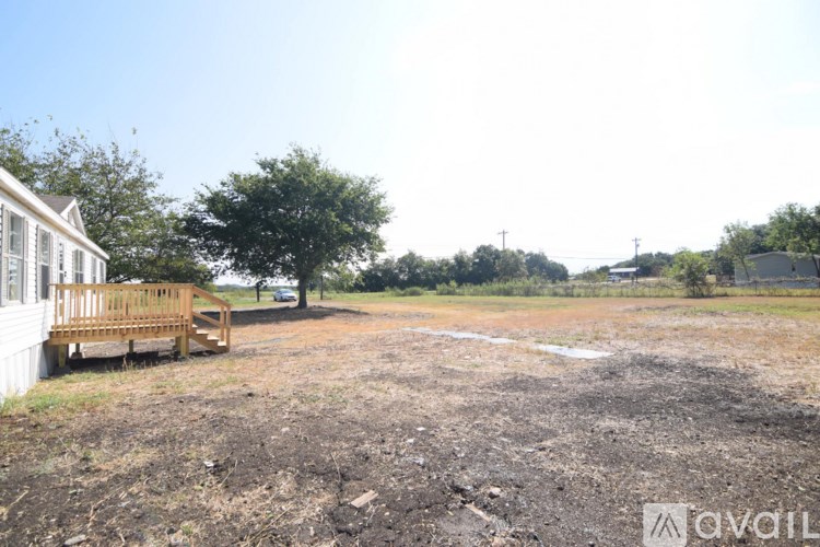 A white house with a wooden deck is situated in a field with a tree and a fence in the background.