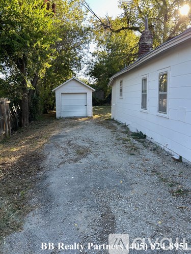 A white house with a garage is situated in a backyard with a gravel driveway.