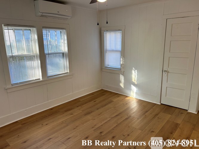 A room with wooden flooring, two windows with blinds, and a white door.