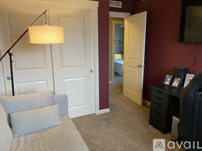 A lamp with a white shade is on a stand in a room with a grey couch and a white door.
