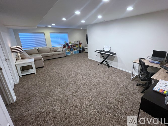 A spacious room with a carpeted floor, a desk with a computer, a couch, a chair, and a small table with a lamp.