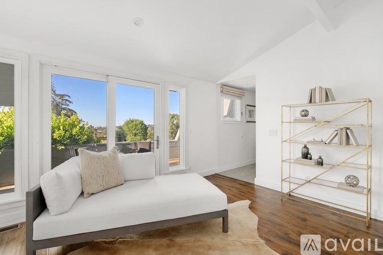 A white sofa with a cushion is in the middle of a room with a large window and a shelf with decorative items.