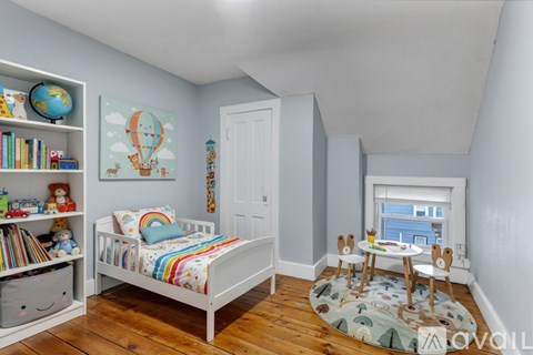 A child's bedroom with a white bed, a bookshelf filled with books, a colorful rug, and a window with white blinds.