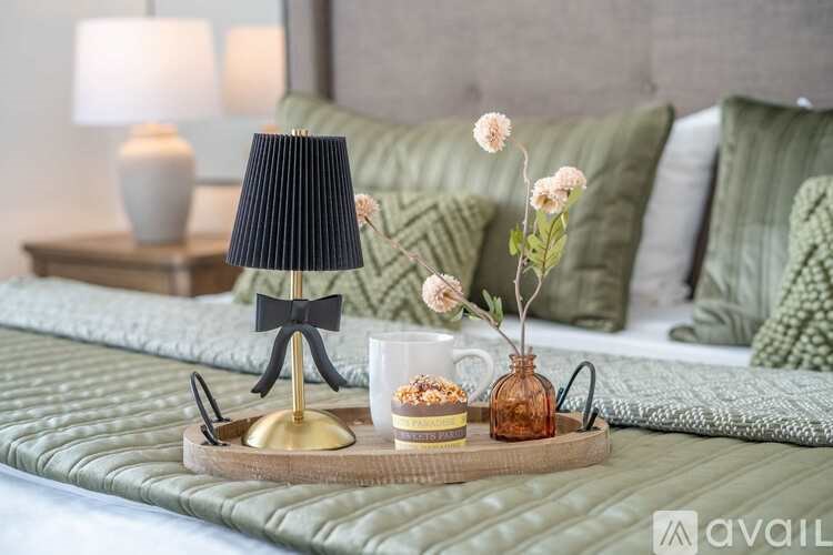 A table with a lamp, a vase with flowers, and a tray with a cup and a cake is in front of a bed with green pillows.