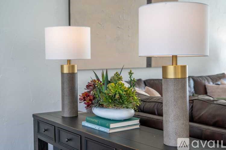 Two table lamps with white shades on a table with a plant and books.