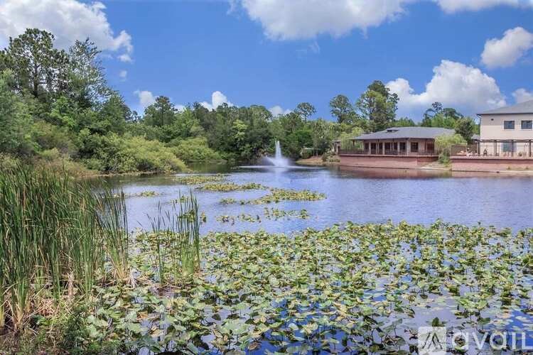 A serene lake with a fountain in the middle, surrounded by lush greenery and a building in the background.