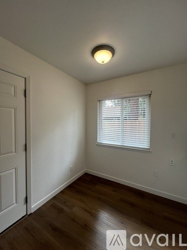 A room with a white door, a window with blinds, and a wooden floor.