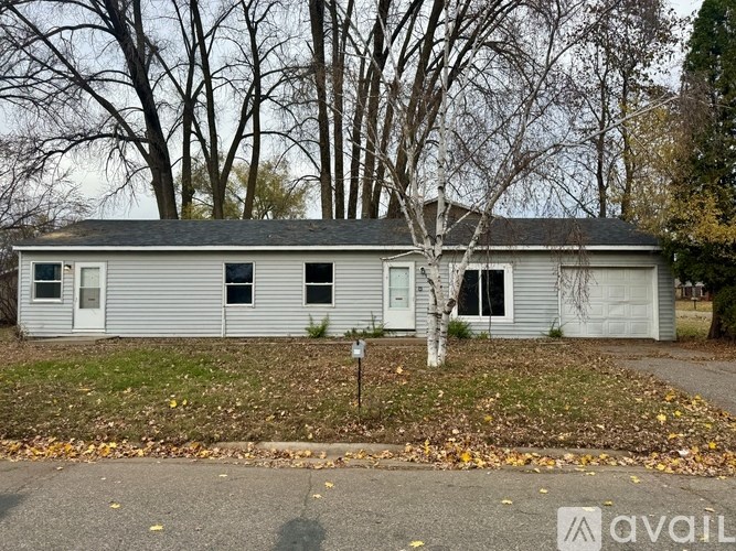 A small house with a grey roof and white siding is surrounded by trees and has a mailbox in front.