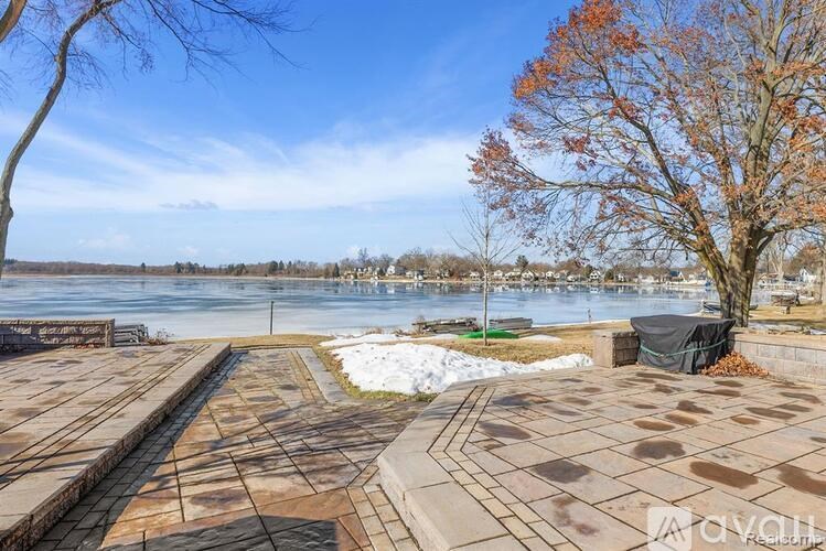 A park with a paved walkway, a tree with orange leaves, and a body of water in the distance.