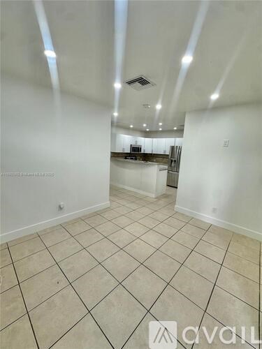 A room with white walls and tiled flooring, with a kitchen area visible in the background.