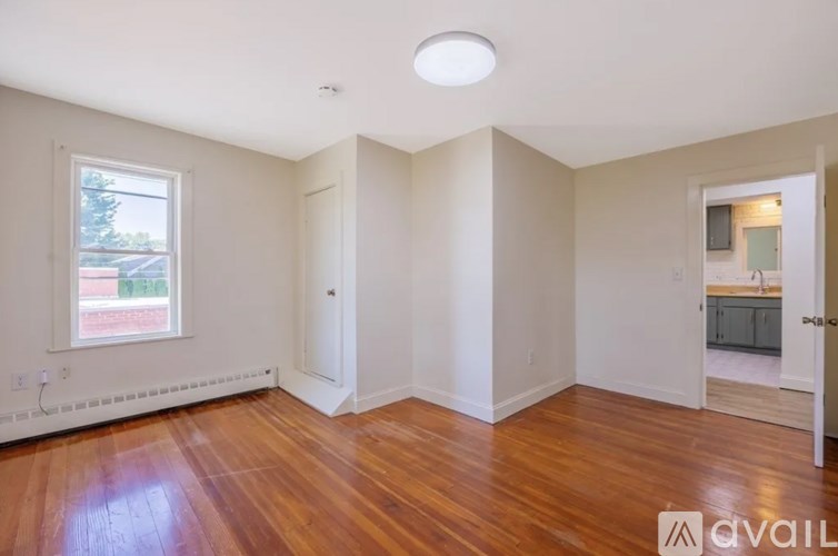 A room with wooden floors and white walls, with a window showing a view of a street and houses.