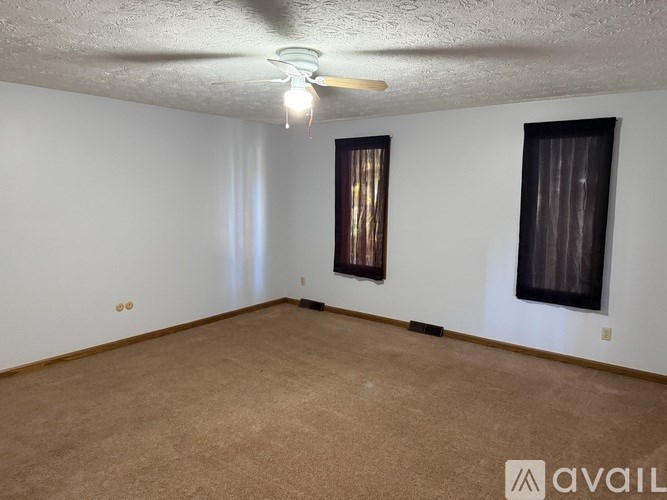 A room with a carpeted floor, a ceiling fan, and two windows with curtains.