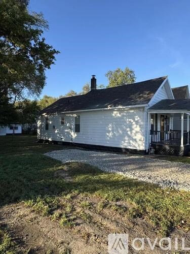 A white house with a black roof and a chimney is surrounded by a gravel driveway and a grassy area.