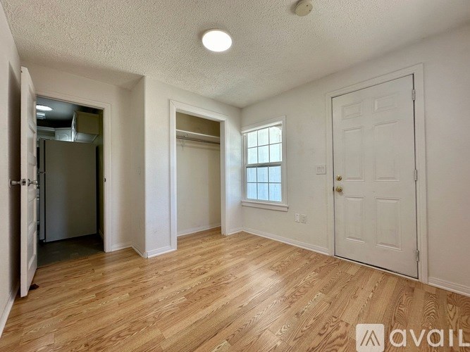A room with wooden flooring and white walls, featuring a refrigerator, a window, and a door.