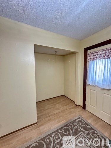 A room with a doorway leading to another room, a window with curtains, and a patterned rug on the floor.