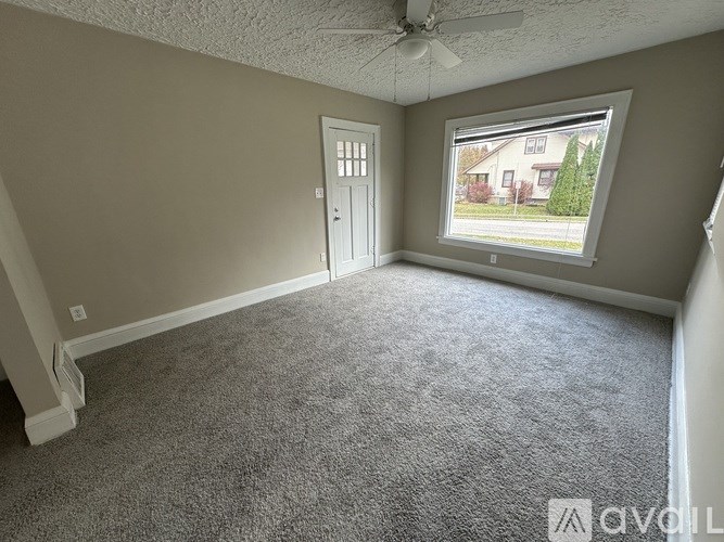 A room with a carpeted floor, a ceiling fan, and a window showing an outdoor view.