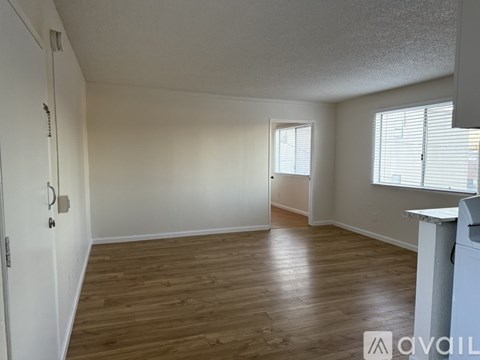 A room with wooden flooring and white walls, with a door on the left and a window on the right.
