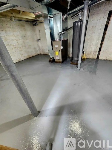 A room with a concrete floor and walls, a metal pipe running along the wall, and a furnace in the corner.