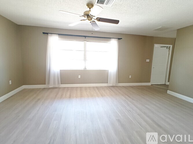 A spacious room with a ceiling fan and light, hardwood floors, and a large window with white curtains.