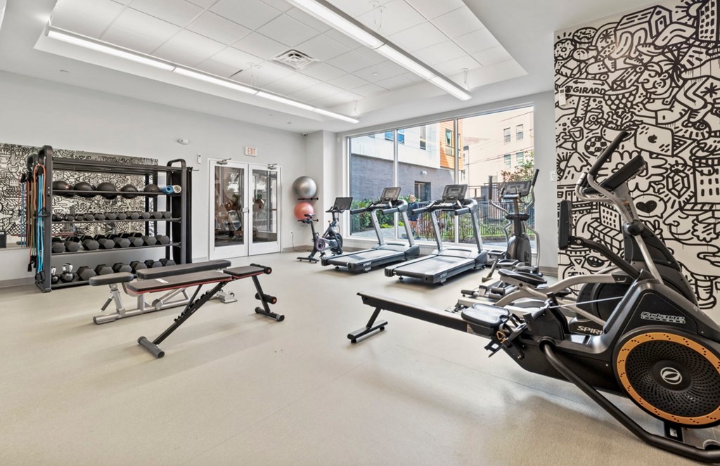A gym with a rowing machine in the foreground and a wall with a black and white pattern on the right.
