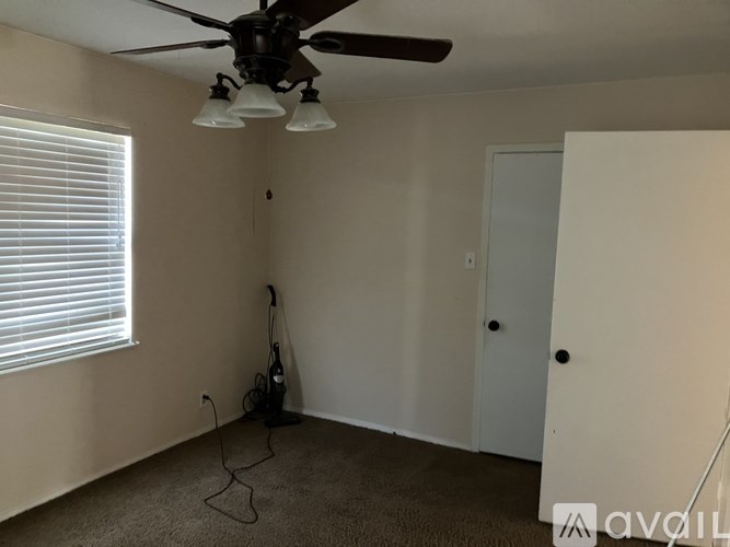 A room with a fan, a window with blinds, a vacuum cleaner, and a door.