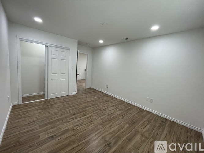 A room with wooden flooring and white walls, with a door on the left and a wall-mounted light fixture.