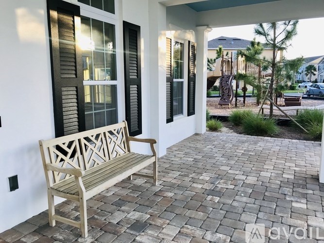 A wooden bench sits on a brick patio in front of a white house with black shutters.