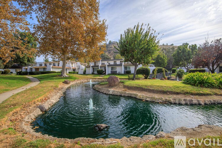 A pond with a fountain in the middle of a grassy area with trees and houses in the background.