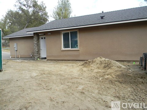 A house with a brown siding and a white door is surrounded by a dirt lot.