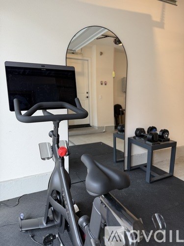 A stationary exercise bike is in a room with a mirror and a table with dumbbells on it.