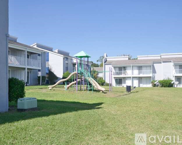 A playground with a green slide in the middle of a grassy area in front of apartment buildings.